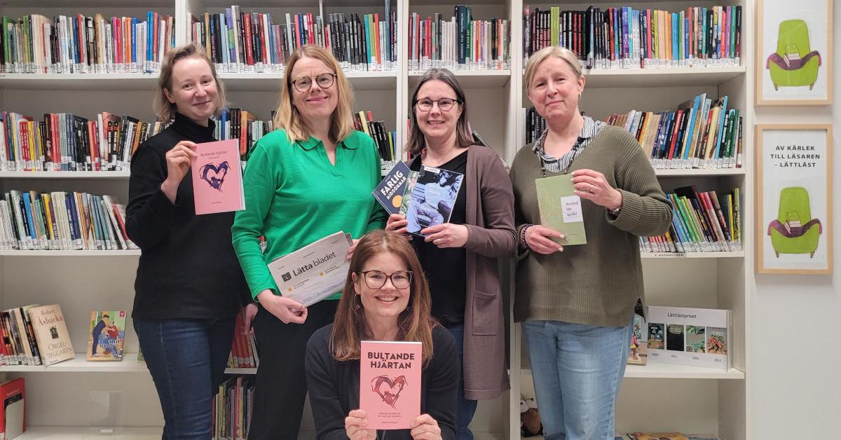 Ett foto av fem glada damer som jobbar på LL-Center. De står framför en bokhylla med lättlästa böcker. I händerna håller de lättalästa böcker, Lätta bladet och boroschyr om lätt språk.